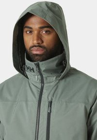 Chaqueta impermeable de color verde claro con cremallera frontal, capucha ajustable y detalle de logo visible en el cuello, que presenta una textura suave.