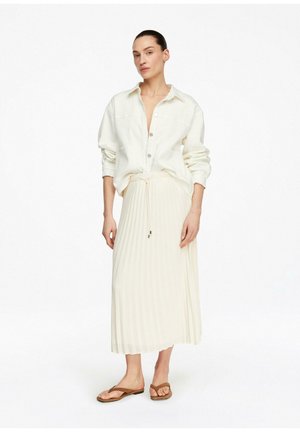 Kobieta ubrana w luźną białą koszulę na guziki, plisowaną kremową spódnicę ze sznurkiem oraz brązowe płaskie sandały, stojąca na białym tle.