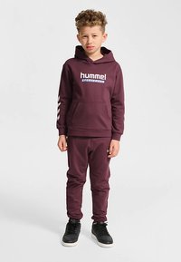 Bordowa bluza z kapturem z białym logo "hummel", wyposażona w kieszeń kangurkę i ściągacze przy rękawach, zestawiona z dopasowanymi bordowymi spodenkami dresowymi i czarnymi trampkami.