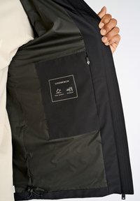 Intérieur de veste noire avec un tissu sombre et lisse, comprenant une poche étiquetée "LINDENBERG" avec des icônes déperlantes et coupe-vent.