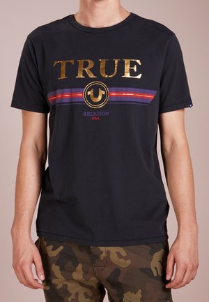 Homme portant un T-shirt noir avec l'inscription dorée "TRUE", une bande violette et rouge et "Religion U.S.A." en dessous, associé à un pantalon camouflage marron-vert.