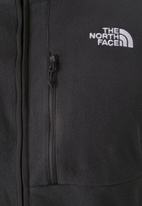 Czarna polarowa kurtka z pionową kieszenią na zamek i białym, wyszywanym logo "THE NORTH FACE" na piersi.