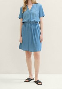 Lichtblauwe denim jurk met korte mouwen, V-hals, elastische tailleband en een zoom die boven de knie eindigt. Gepaarde met zwarte platte sandalen.