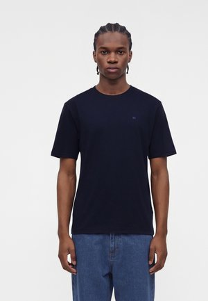 Ung mand med fletninger står med fronten mod kameraet iført en ensfarvet sort kortærmet T-shirt og blå jeans mod en hvid baggrund.