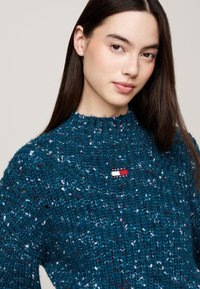 Suéter grueso de color azul verdoso con cuello alto y patrón texturizado, que presenta un pequeño parche rectangular con el logo "TOMMY JEANS" en el pecho.