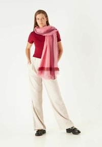 Écharpe rose clair avec une texture subtile et des bords frangés, superposée à un haut rouge profond, associée à un pantalon beige large et des chaussures noires.