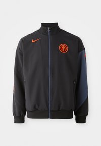 Crna sportska jakna s visokim ovratnikom, narančastim detaljima, zatvaračem i bočnim džepovima. Ima logo na prsima i kontrastne rukave.