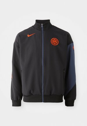 Giacca sportiva nera con colletto alto, dettagli arancioni, chiusura con zip e tasche laterali. Presenta un logo sul petto e maniche a contrasto.