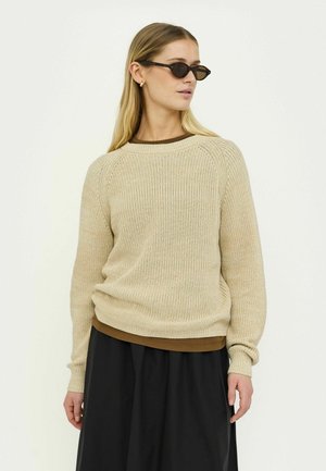 Maglione beige lavorato a maglia con trama a coste, maniche raglan e orlo marrone. Abbinato a una gonna nera e occhiali da sole scuri.