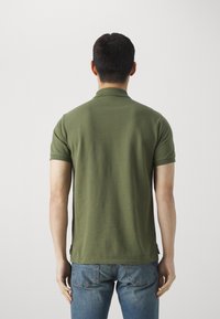 Polo verde con colletto classico, maniche corte e una texture liscia, abbinato a jeans blu. Slit laterali visibili all'orlo inferiore.