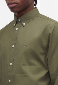 Gros plan d'une personne portant une chemise boutonnée vert olive avec un col boutonné et un petit logo rouge, blanc et bleu sur la poitrine.