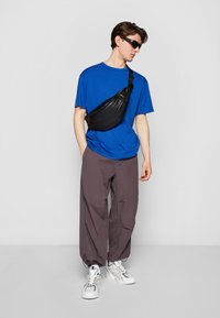 Sac en cuir noir porté en bandoulière sur un t-shirt bleu, associé à un pantalon baggy gris et des baskets blanches. Le modèle porte des lunettes de soleil noires.