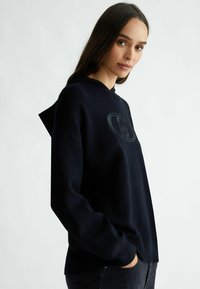 Felpa blu navy con tasca a marsupio, polsini a costine e cappuccio. Presenta un logo discreto sul petto. Il tessuto appare testurizzato e morbido.
