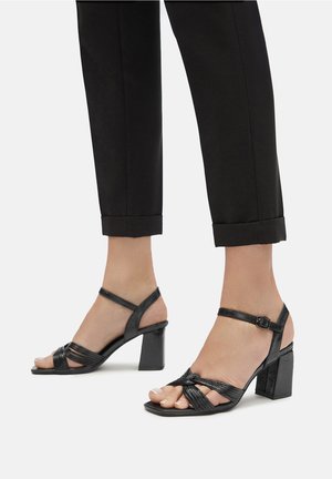 Fekete sarokszandál, keresztezett pántokkal, nyitott orral és négyzetes orrformával. Állítható bokapánt; tömbsarok.