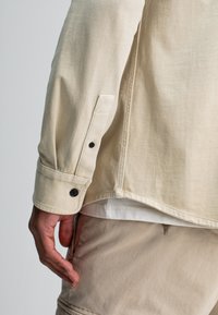 Close-up van de hand van een persoon die naast zijn lichaam rust, gekleed in een beige langemouwen shirt met zwarte knopen en lichtbeige broek.