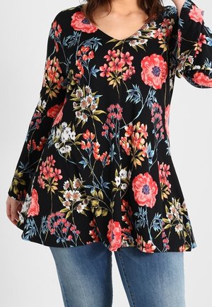 Woman wearing a black long-sleeve tunic with red, white, and blue floral print paired with blue jeans, cropped at shoulders and thighs.