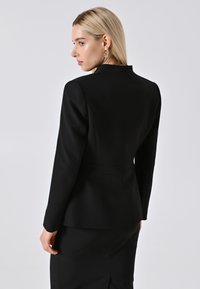 Donna bionda in blazer aderente nero e gonna, che indossa grandi orecchini a cerchio dorati, rivolta a sinistra su uno sfondo grigio neutro.
