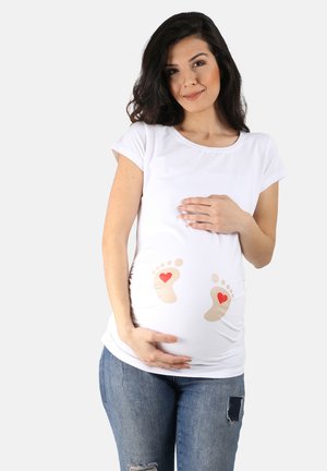 Weißes Maternity-T-Shirt mit kurzen Ärmeln, auf der Vorderseite mit beigen Babyfüßen und roten Herzen bedruckt, kombiniert mit lässigen blauen Jeans.