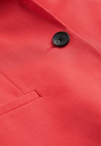 Nahaufnahme von rotem Stoff mit glatter Textur, einem einzelnen schwarzen Knopf mit vier Löchern und einer genähten Taschen-Detail.