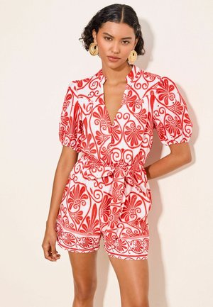 Vrouw die een rood-wit patroon korte mouwen playsuit draagt met een strik in de taille en grote ronde oorbellen, staat tegen een effen achtergrond.