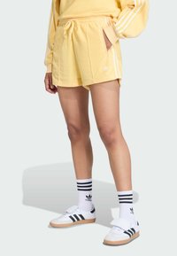 Žlté bavlnené šortky s bielymi pruhmi, bočnými vreckami a vyšitým logom. Skombinované s čierno-bielymi pruhovanými ponožkami a teniskami.