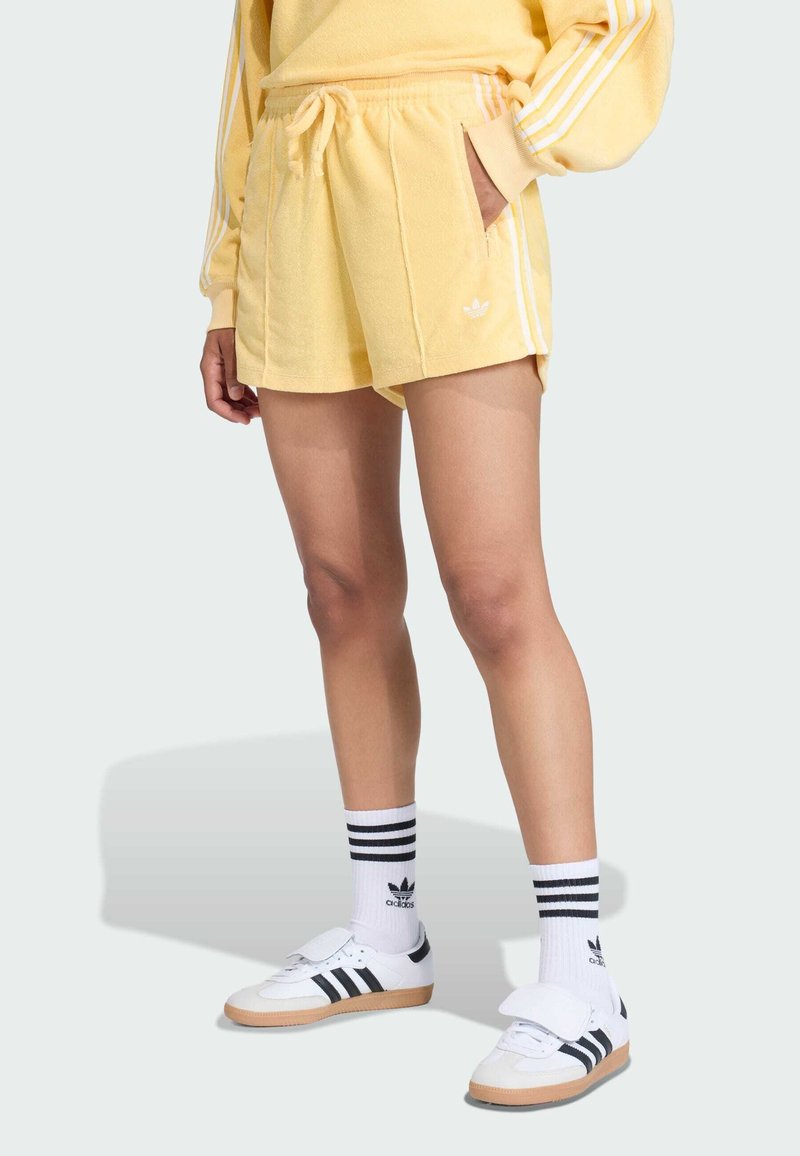 Žlté bavlnené šortky s bielymi pruhmi, bočnými vreckami a vyšitým logom. Skombinované s čierno-bielymi pruhovanými ponožkami a teniskami.