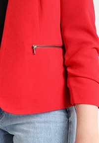 Blazer rouge en tissu texturé, avec une poche zippée argentée sur le côté inférieur droit et des manches légèrement retroussées.