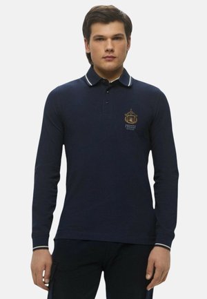 Polo a maniche lunghe colore blu navy con colletto, caratterizzato da accenti a strisce bianche e un emblema dorato sul petto. Realizzato in tessuto di misto cotone liscio.