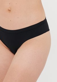 Woman wearing black mid-rise bikini briefs, showing lower abdomen and upper thighs against a plain background.