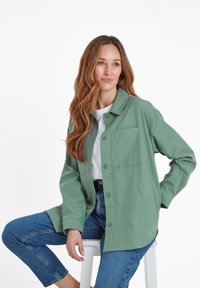 Green cotton jacket with a button-up front, pointed collar, and a chest pocket, paired with blue jeans, sitting against a white background.