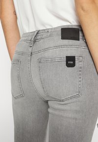 Primer plano de una persona que lleva pantalones vaqueros gris claro con bolsillos traseros, parches de marca negra y una camiseta blanca metida por dentro.