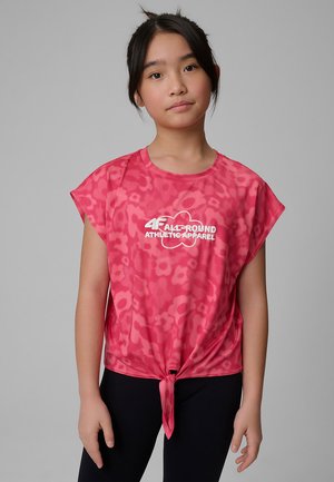 Joven chica con cabello oscuro que lleva una camiseta deportiva rosa con estampado, logo y nudo frontal, combinada con mallas negras, de pie frente a un fondo gris.