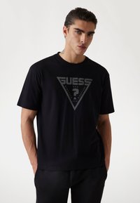 Juodas medvilnės marškinėliai su dideliu pilku trikampiu logotipu, kuriame parašyta "GUESS", "U.S.A." ir numeriai, trumpomis rankovėmis ir apvalia iškirpte.