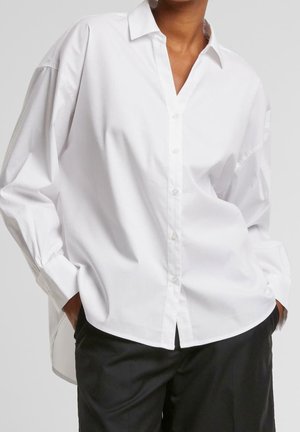 Osoba ubrana w oversize'ową białą koszulę na guziki z kołnierzem, czarne spodnie, stoi z rękami w kieszeniach na tle jednolitego tła.