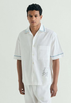 Joven que lleva una camisa blanca de manga corta con botones y costuras azules, con el texto "I [heart] PR" en la zona del bolsillo delantero.