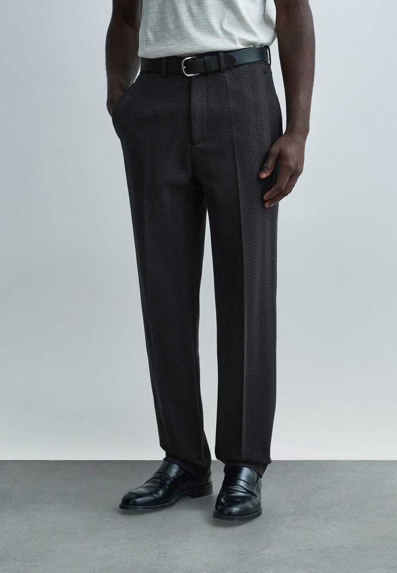 Μαύροι παντελόνες ψαροκόκαλο με χαλαρή εφαρμογή, θηλιές για ζώνη με μαύρη ζώνη και συνδυάζονται με γυαλιστερά μαύρα δερμάτινα παπούτσια.