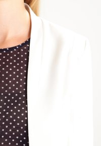 Femme portant une chemise noire à motifs d'étoiles blanches et un blazer blanc, montrée du cou à l'épaule sur fond blanc.