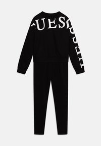 Svart sweatshirt- och byxset, tillverkat av bomull. Sweatshirten har en stor vit "GUESS"-logotyp på framsidan och ärmarna. Slät textur.