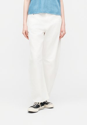 Woman wearing white loose-fit trousers, black and white sneakers, and a cropped light blue top against a plain white background.