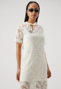 Robe en dentelle de couleur blanc cassé, à manches courtes et avec des franges, dotée d'un décolleté en forme de clé et de motifs floraux tout au long.