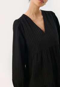 Robe noire avec un décolleté en V, en tissu texturé, manches bouffantes longues et taille froncée. Elle présente des rayures horizontales subtiles et une silhouette fluide.