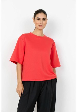 Ženska s temnimi lasmi, vezanimi nazaj, oblečena v ohlapno rdečo majico s kratkimi rokavi in črne hlače, stoji z eno roko v žepu na belem ozadju.