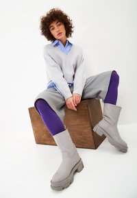 Suéter gris, camisa azul, pantalones cortos grises de pierna ancha, leggings morados, botas gris a medio pantorrilla con suelas gruesas, sentado en una caja de madera.