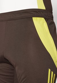 Pantalon de sport marron avec un panneau latéral en mesh jaune, doté d'une taille élastique et de trois bandes verticales sur la couture extérieure.