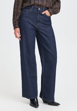 Persona che indossa jeans blu scuro a gamba larga, una camicia a quadri marrone e stivali neri con punta appuntita, in piedi con una mano in tasca.