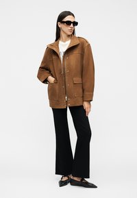 Femme portant une veste marron, un t-shirt blanc, un pantalon noir à jambes larges, des lunettes de soleil noires et des ballerines noires pointues, debout avec une main dans la poche.