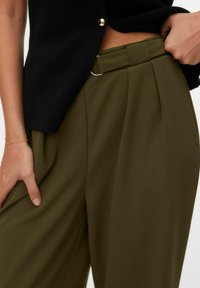 Persoon draagt olijfgroene broek met hoge taille en ceintuurdetail, gecombineerd met een cropped zwart topje met knopen, met blote buik en hand op heup.