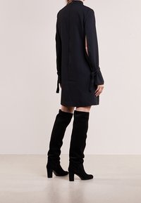 Robe noire à manches longues avec fentes sur les côtés et accents de manches nouées, portée avec des bottes en suède noires à hauteur de genou sur une surface claire.