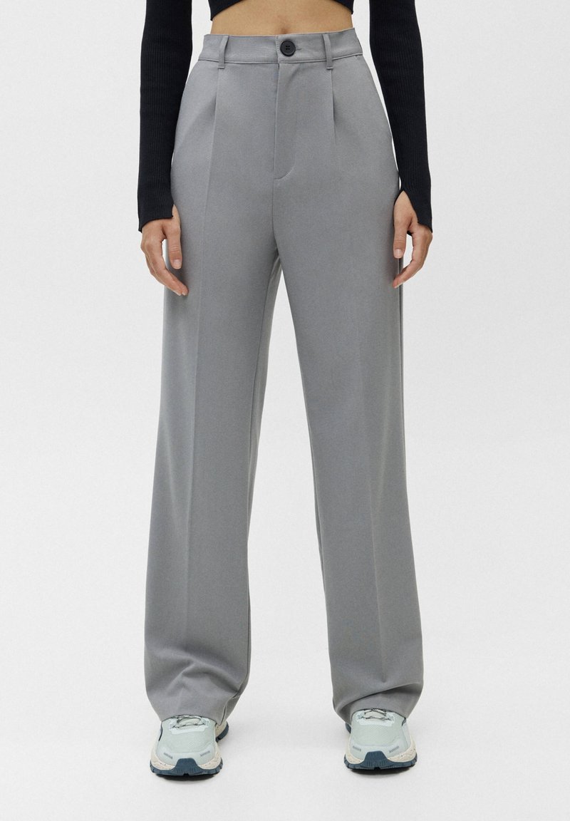 Femme portant un pantalon gris taille haute à jambes larges avec des plis devant et un top court noir à manches longues, assorti à des baskets blanches et grises.