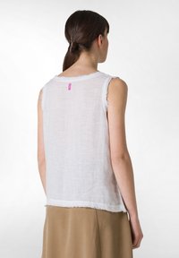 Top sin mangas de lino blanco con bordes deshilachados, escote redondeado y una pequeña etiqueta rosa en la parte posterior. Combinado con una falda beige.